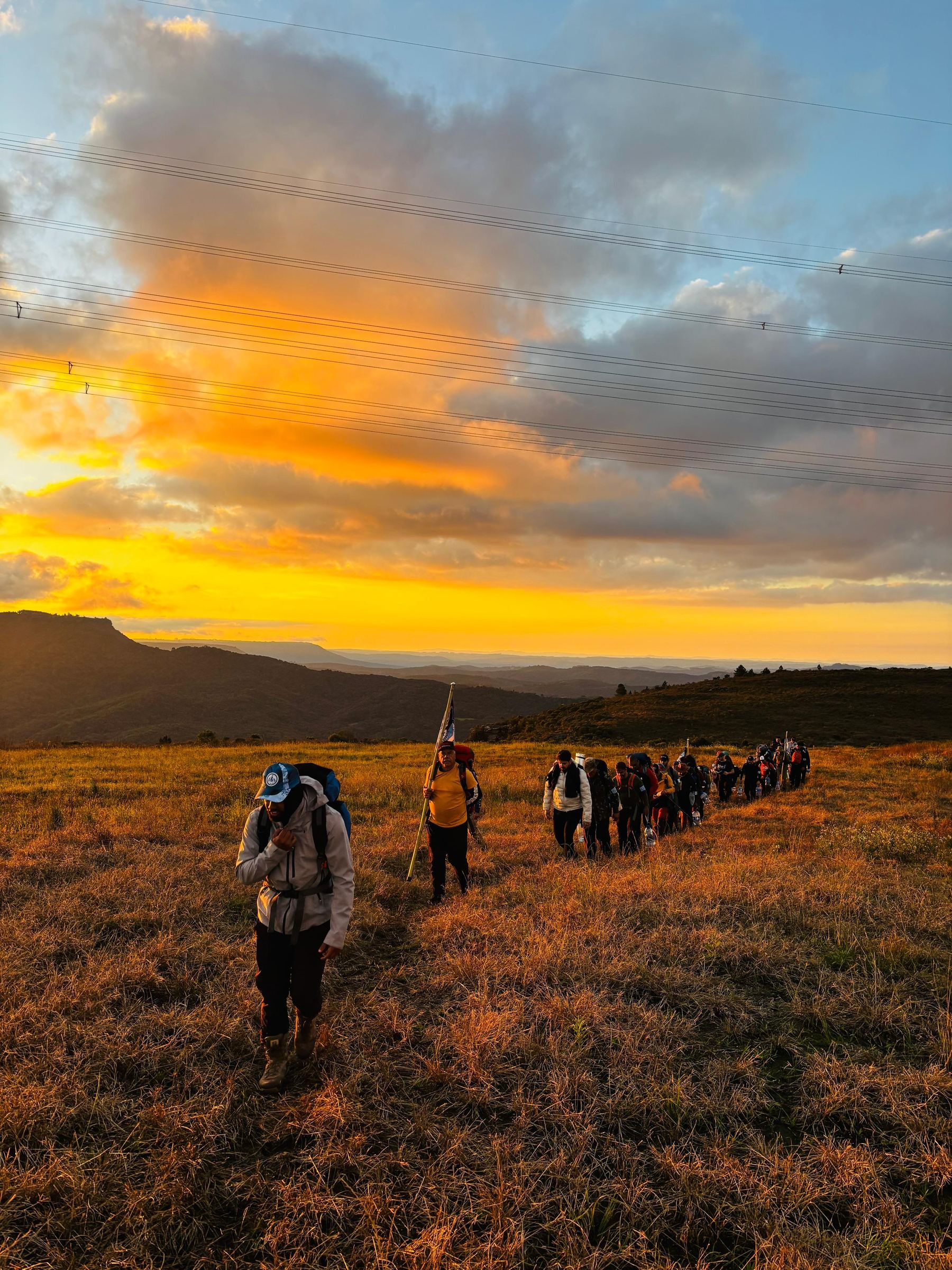 Fila de homens com mochilas caminhando por um campo aberto ao pôr do sol